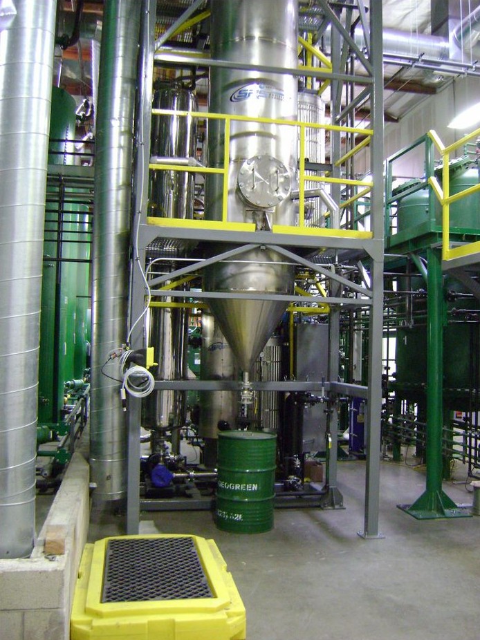 Stainless-steel process vessel on a steel platform with yellow safety railings, inside a processing hall